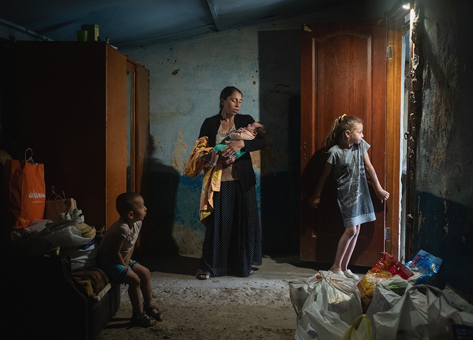 Une femme tient un bébé dans ses bras dans une chambre; à sa droite un jeune enfant la regarde et à sa gauche une jeune fille se tient à côté de la porte d'entrée.