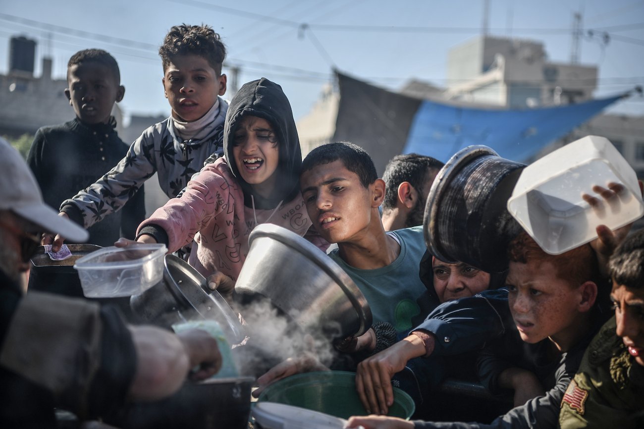 Un groupe de jeunes enfants en état de détresse tiennent entre les mains des récipients qu'ils tendent vers un individu pour pouvoir recevoir de la nourriture 